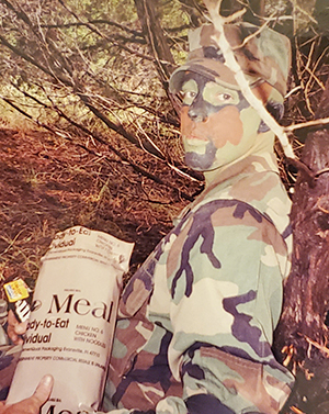 Photo of Brandon Womack dressed in Air Force camouflage eating MRE in forest.